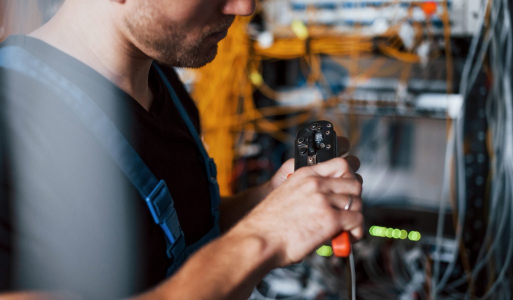 Young man in uniform works with internet equipment and wires in server room.